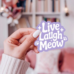 May include: A lilac sticker featuring the text "Live Laugh Meow" in white, with paw prints and star details. The sticker is held in a hand with a white manicure, set against a soft-focus backdrop.