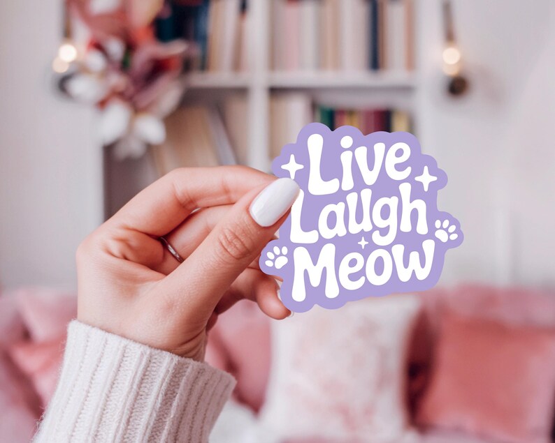 May include: A lilac sticker featuring the text "Live Laugh Meow" in white, with paw prints and star details. The sticker is held in a hand with a white manicure, set against a soft-focus backdrop.