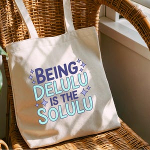 May include: Cream-colored tote bag with the text "BEING DELULU IS THE SOLULU" in blue and light blue. The bag has two handles and is sitting on a wicker chair.