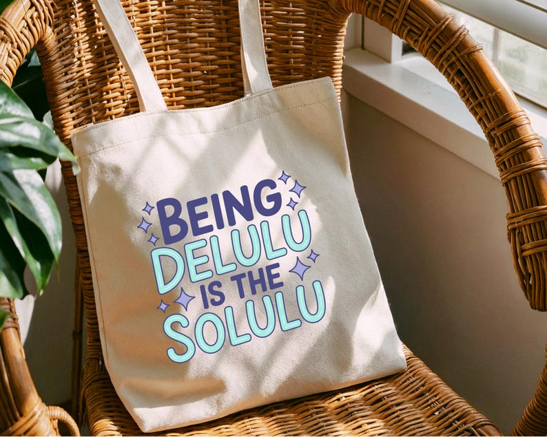 May include: Cream-colored tote bag with the text "BEING DELULU IS THE SOLULU" in blue and light blue. The bag has two handles and is sitting on a wicker chair.