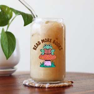 May include: A clear glass tumbler, shaped like a can, filled with a light-coloured drink and ice. The glass has a colourful graphic of a frog reading a book on a mushroom, with the text "Read More Books". A reusable straw is inserted.