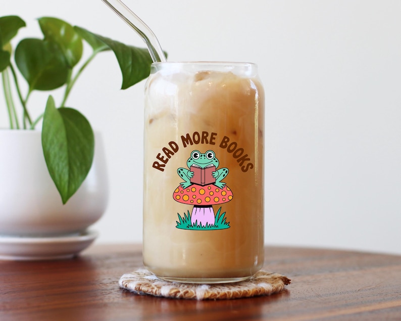 May include: A clear glass tumbler, shaped like a can, filled with a light-coloured drink and ice. The glass has a colourful graphic of a frog reading a book on a mushroom, with the text "Read More Books". A reusable straw is inserted.