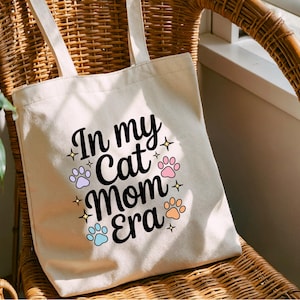 May include: A cream-coloured tote bag with the text "In my Cat Mom Era" in black script, surrounded by paw prints and stars in pastel colours. The bag has two cream-coloured handles and is sitting on a wicker chair.