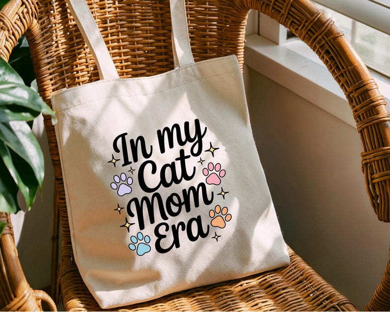 May include: A cream-coloured tote bag with the text "In my Cat Mom Era" in black script, surrounded by paw prints and stars in pastel colours. The bag has two cream-coloured handles and is sitting on a wicker chair.