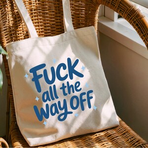 K&ouml;nnte beinhalten: Eine cremefarbene Tragetasche mit dem Text "Fuck all the way off" in blauer Schrift. Die Tasche hat zwei cremefarbene Tr&auml;ger und liegt auf einem Korbstuhl.