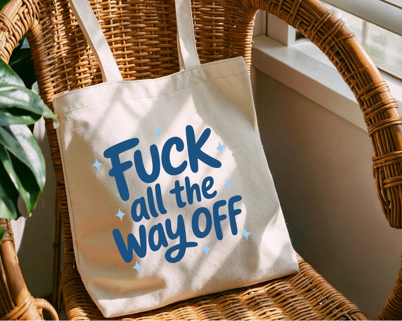 K&ouml;nnte beinhalten: Eine cremefarbene Tragetasche mit dem Text "Fuck all the way off" in blauer Schrift. Die Tasche hat zwei cremefarbene Tr&auml;ger und liegt auf einem Korbstuhl.