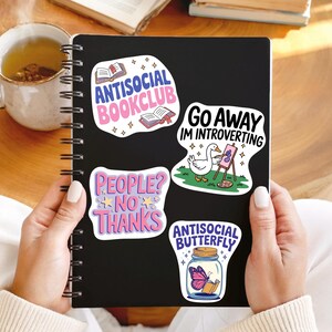 K&ouml;nnte beinhalten: Ein schwarzes Notizbuch mit vier bunten Aufklebern. Die Aufkleber zeigen S&auml;tze wie "Antisocial Bookclub" und "Go Away I'm Introverting". Eine Tasse Tee und B&uuml;cher sind im Hintergrund zu sehen. Das Notizbuch wird von einer Person gehalten.