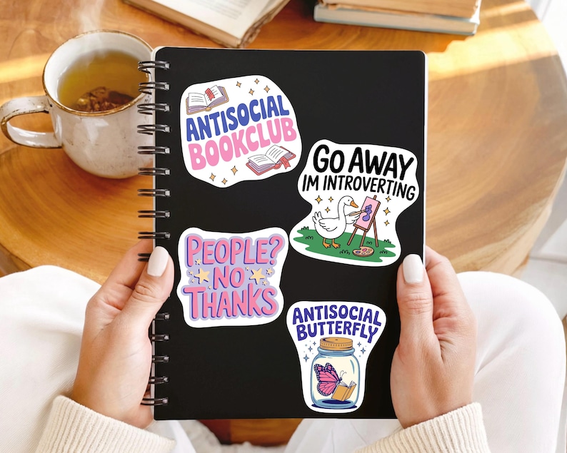 K&ouml;nnte beinhalten: Ein schwarzes Notizbuch mit vier bunten Aufklebern. Die Aufkleber zeigen S&auml;tze wie "Antisocial Bookclub" und "Go Away I'm Introverting". Eine Tasse Tee und B&uuml;cher sind im Hintergrund zu sehen. Das Notizbuch wird von einer Person gehalten.
