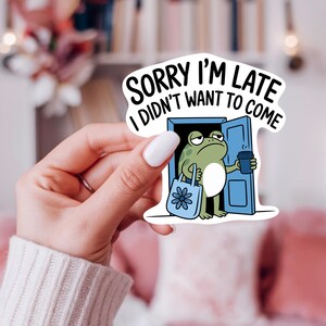 K&ouml;nnte beinhalten: Ein wei&szlig;er Aufkleber mit dem Text "SORRY I'M LATE I DIDN'T WANT TO COME". Der Aufkleber zeigt einen Cartoon-Frosch, der eine Kaffeetasse und eine Tragetasche h&auml;lt und in einer blauen T&uuml;r steht. Der Frosch ist gr&uuml;n und hat einen m&uuml;rrischen Gesichtsausdruck.