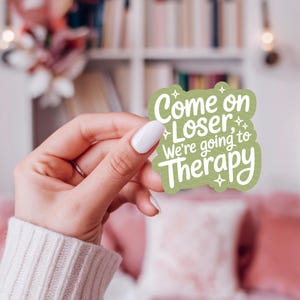 以下が含まれることがあります： 「Come on Loser, We're going to Therapy」の白文字が入った緑色のステッカー。白いマニキュアを塗った手で持っています。背景には本棚とピンクのソファがあります。