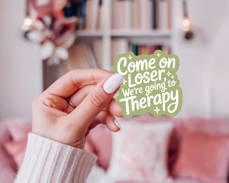 以下が含まれることがあります： 「Come on Loser, We're going to Therapy」の白文字が入った緑色のステッカー。白いマニキュアを塗った手で持っています。背景には本棚とピンクのソファがあります。