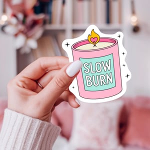 May include: A pink candle sticker with a light blue label that reads "SLOW BURN" in white text. The candle has a yellow flame with a heart shape in the middle. Black star accents surround the sticker.