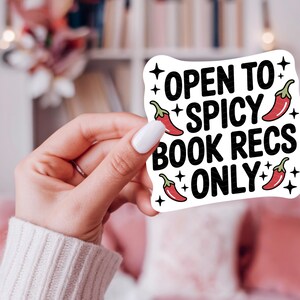 May include: A white sticker with the text "OPEN TO SPICY BOOK RECS ONLY" in black, with red chili pepper illustrations. The sticker is held by a person with a white sweater and white nail polish.
