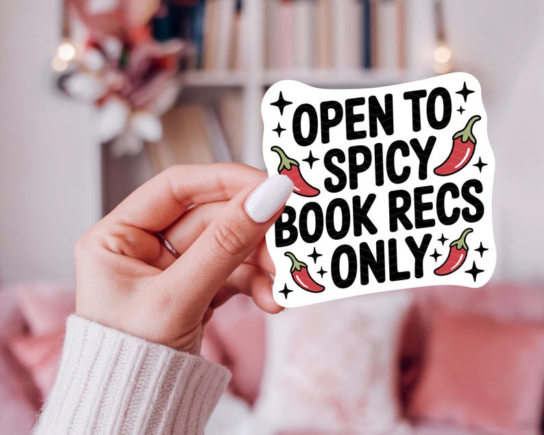 May include: A white sticker with the text "OPEN TO SPICY BOOK RECS ONLY" in black, with red chili pepper illustrations. The sticker is held by a person with a white sweater and white nail polish.