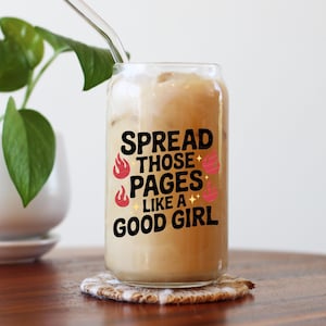May include: A clear glass tumbler filled with a creamy beverage and ice, featuring the text "SPREAD THOSE PAGES LIKE A GOOD GIRL" in black, with red flame and pink dumbbell graphics. A glass straw is inserted. The tumbler sits on a patterned coaster.