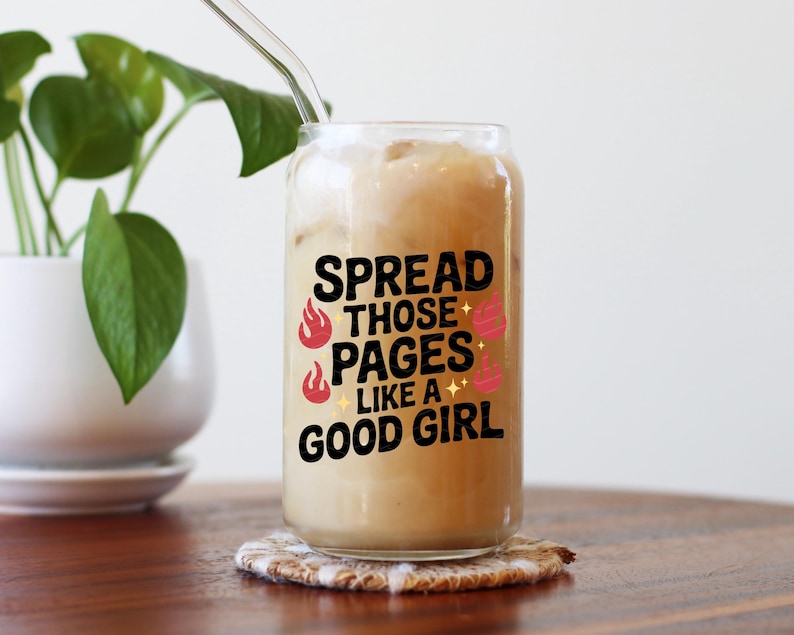 May include: A clear glass tumbler filled with a creamy beverage and ice, featuring the text "SPREAD THOSE PAGES LIKE A GOOD GIRL" in black, with red flame and pink dumbbell graphics. A glass straw is inserted. The tumbler sits on a patterned coaster.