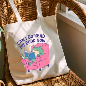 K&ouml;nnte beinhalten: Cremefarbene Tragetasche mit dem Text "CAN I GO READ MY BOOK NOW". Die Tasche zeigt einen Cartoon-Frosch, der ein Buch liest, w&auml;hrend er in einem rosa Sessel sitzt. Die Tasche hat zwei cremefarbene Tr&auml;ger und sitzt auf einem Korbstuhl.