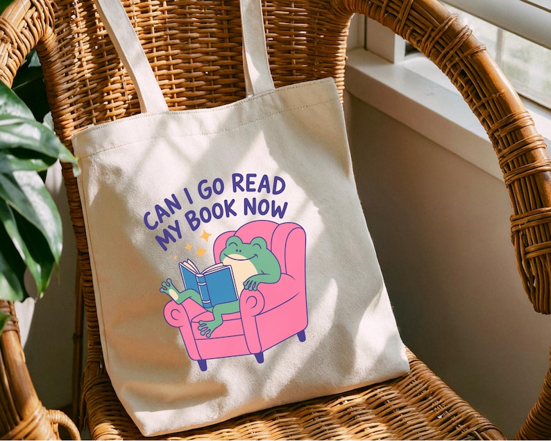 K&ouml;nnte beinhalten: Cremefarbene Tragetasche mit dem Text "CAN I GO READ MY BOOK NOW". Die Tasche zeigt einen Cartoon-Frosch, der ein Buch liest, w&auml;hrend er in einem rosa Sessel sitzt. Die Tasche hat zwei cremefarbene Tr&auml;ger und sitzt auf einem Korbstuhl.
