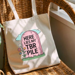 May include: A cream-coloured tote bag featuring a pink tombstone design with the text "HERE LIES MY TBR PILE". The graphic includes a blue cloud, flowers, and green grass. The bag has two cream-coloured handles.