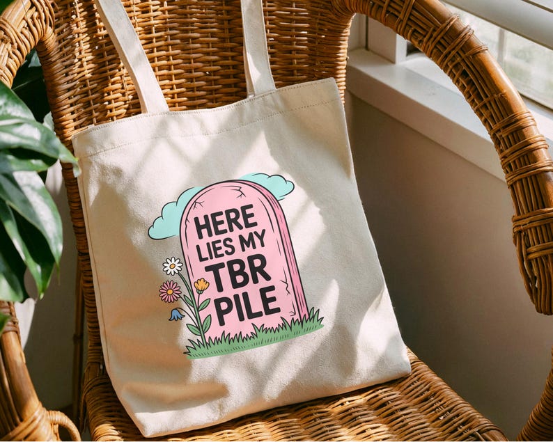 May include: A cream-coloured tote bag featuring a pink tombstone design with the text "HERE LIES MY TBR PILE". The graphic includes a blue cloud, flowers, and green grass. The bag has two cream-coloured handles.