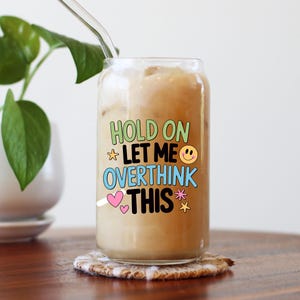 May include: A clear glass can-shaped tumbler filled with a light brown beverage and ice. The tumbler has the words "HOLD ON LET ME OVERTHINK THIS" in colorful text. A clear glass straw is inserted into the drink, and the glass sits on a patterned coaster.