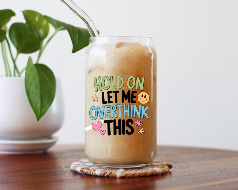 May include: A clear glass can-shaped tumbler filled with a light brown beverage and ice. The tumbler has the words "HOLD ON LET ME OVERTHINK THIS" in colorful text. A clear glass straw is inserted into the drink, and the glass sits on a patterned coaster.
