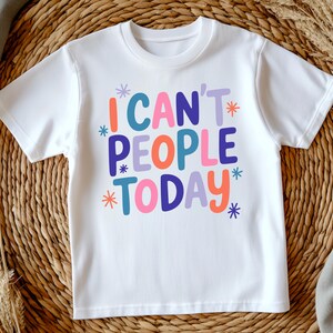 K&ouml;nnte beinhalten: Wei&szlig;es T-Shirt mit dem Schriftzug "I CAN'T PEOPLE TODAY" in farbenfrohen, verspielten Buchstaben. Der Text ist in verschiedenen Farben gehalten, darunter T&uuml;rkis, Orange, Lila, Pink und Blau, mit Sternakzenten. Das Shirt wird auf einem gewebten Hintergrund pr&auml;sentiert.