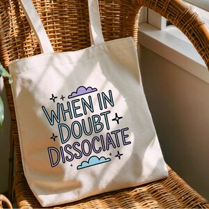以下が含まれることがあります： クリーム色のトートバッグ。テキスト「WHEN IN DOUBT DISSOCIATE」が青と紫で書かれています。バッグには2つのハンドルがあり、籐の椅子の上に置かれています。テキストは星と雲で囲まれています。