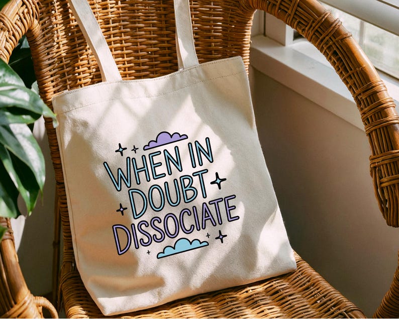 以下が含まれることがあります： クリーム色のトートバッグ。テキスト「WHEN IN DOUBT DISSOCIATE」が青と紫で書かれています。バッグには2つのハンドルがあり、籐の椅子の上に置かれています。テキストは星と雲で囲まれています。