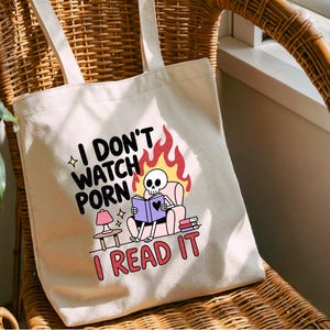 May include: Cream-colored tote bag with a humorous graphic. The design features a skeleton reading a book in a chair, with flames and text that reads "I DON'T WATCH PORN I READ IT". The bag has two handles.