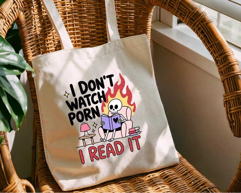 May include: Cream-colored tote bag with a humorous graphic. The design features a skeleton reading a book in a chair, with flames and text that reads "I DON'T WATCH PORN I READ IT". The bag has two handles.