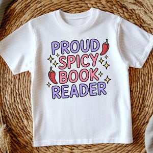 May include: White t-shirt with the text "PROUD SPICY BOOK READER" in purple and pink bubble letters. The design includes chili pepper illustrations and star accents. The shirt is displayed on a woven background.