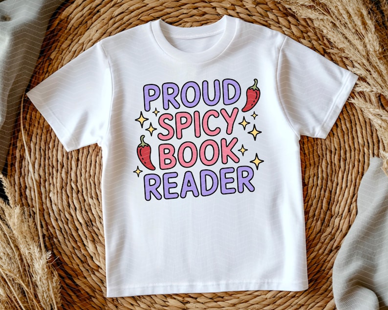May include: White t-shirt with the text "PROUD SPICY BOOK READER" in purple and pink bubble letters. The design includes chili pepper illustrations and star accents. The shirt is displayed on a woven background.