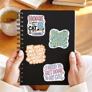 May include: A black notebook with spiral binding, adorned with four cat-themed stickers. The stickers display phrases such as "Home is where the cat is welcome" and "Proud Cat Mom." A mug of tea is visible.