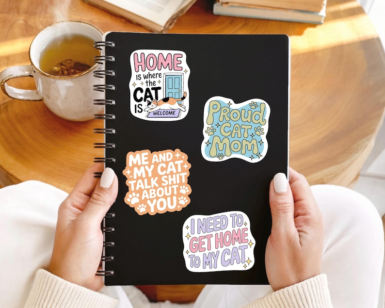 May include: A black notebook with spiral binding, adorned with four cat-themed stickers. The stickers display phrases such as "Home is where the cat is welcome" and "Proud Cat Mom." A mug of tea is visible.