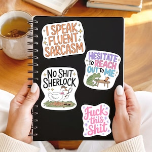 May include: A black notebook with four humorous stickers. The stickers feature phrases like "I Speak Fluent Sarcasm," "No Shit Sherlock," "Hesitate to Reach Out to Me," and "Fuck This Shit." The notebook is held by a person.