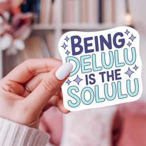 May include: A white sticker with the text "Being Delulu Is The Solulu" in blue and teal, with star accents. The sticker is held by a hand with white nail polish, against a blurred background.