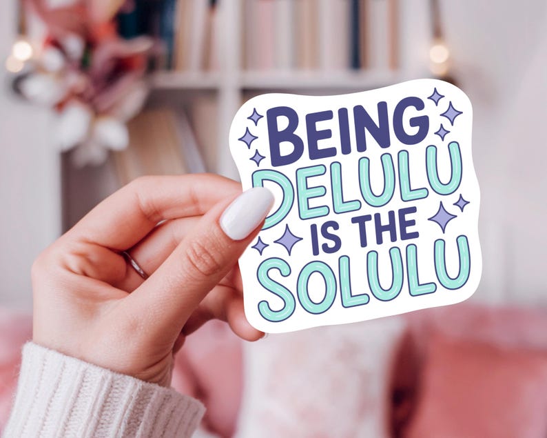 May include: A white sticker with the text "Being Delulu Is The Solulu" in blue and teal, with star accents. The sticker is held by a hand with white nail polish, against a blurred background.