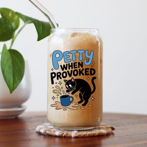 K&ouml;nnte beinhalten: Ein Glas Eiskaffee mit einem durchsichtigen Strohhalm, auf einem Untersetzer. Das Glas zeigt ein Cartoon-Design einer schwarzen Katze, eine versch&uuml;ttete blaue Tasse und den Text "Petty When Provoked" in Blau und Wei&szlig;.