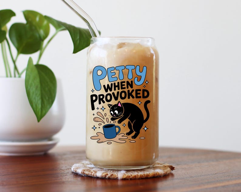 K&ouml;nnte beinhalten: Ein Glas Eiskaffee mit einem durchsichtigen Strohhalm, auf einem Untersetzer. Das Glas zeigt ein Cartoon-Design einer schwarzen Katze, eine versch&uuml;ttete blaue Tasse und den Text "Petty When Provoked" in Blau und Wei&szlig;.
