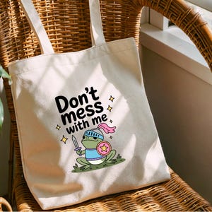 May include: Cream-colored tote bag with the text "Don't mess with me" and a cartoon frog warrior illustration. The frog is green, wearing a helmet, holding a sword and shield. The bag has two long handles.