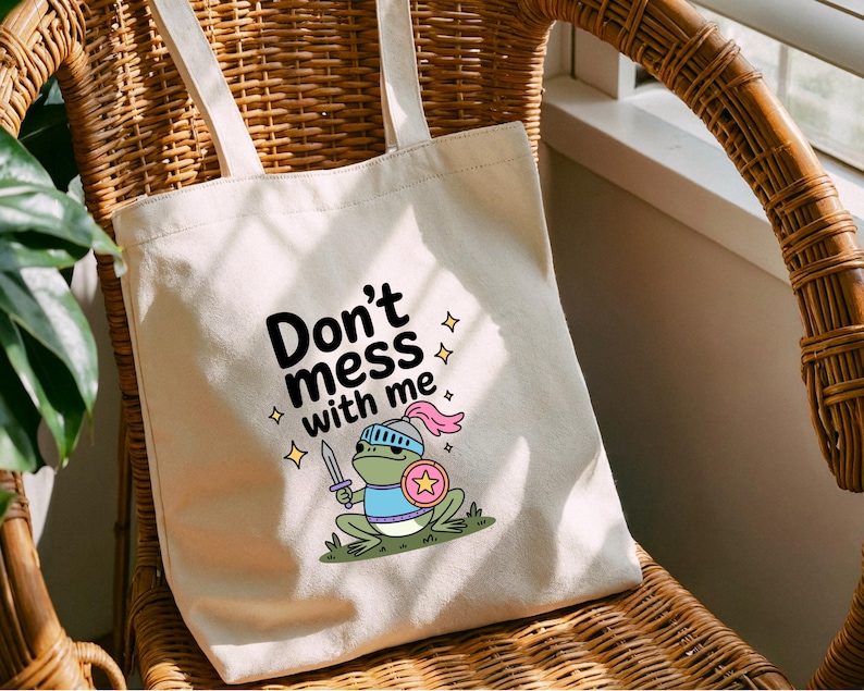 May include: Cream-colored tote bag with the text "Don't mess with me" and a cartoon frog warrior illustration. The frog is green, wearing a helmet, holding a sword and shield. The bag has two long handles.