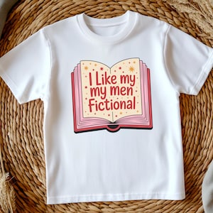 May include: White t-shirt with a graphic of an open book. The book design has a pink border and gold star accents, and the text reads "I Like my men Fictional" in red and pink.