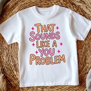 May include: White t-shirt with the phrase "THAT SOUNDS LIKE A YOU PROBLEM" in orange and pink bubble letters. Pink starburst graphics are placed around the text. The shirt is displayed on a woven surface.