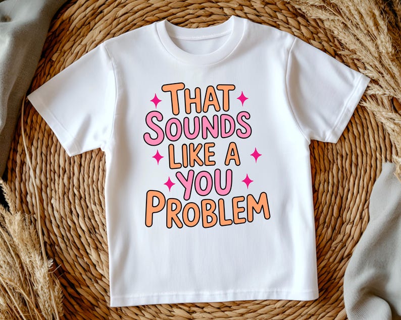 May include: White t-shirt with the phrase "THAT SOUNDS LIKE A YOU PROBLEM" in orange and pink bubble letters. Pink starburst graphics are placed around the text. The shirt is displayed on a woven surface.