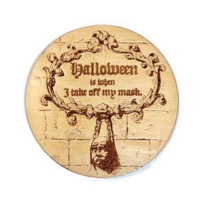 May include: A round wooden coaster with a laser-engraved design. The design features a decorative frame with the text "Halloween is when I take off my mask."