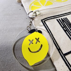 K&ouml;nnte beinhalten: Ein klarer Acryl-Schl&uuml;sselanh&auml;nger mit einem gelben Zitronen-Design. Die Zitrone hat schwarze kreuzf&ouml;rmige Augen und ein gebogenes L&auml;cheln. Der Schl&uuml;sselanh&auml;nger ist an einer silbernen Kette und einem wei&szlig;en Rei&szlig;verschlussanh&auml;nger befestigt. Im Hintergrund befindet sich eine Leinwandtasche mit einem Zitronenscheiben-Design.