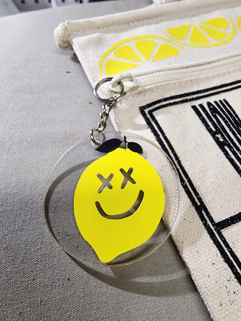 K&ouml;nnte beinhalten: Ein klarer Acryl-Schl&uuml;sselanh&auml;nger mit einem gelben Zitronen-Design. Die Zitrone hat schwarze kreuzf&ouml;rmige Augen und ein gebogenes L&auml;cheln. Der Schl&uuml;sselanh&auml;nger ist an einer silbernen Kette und einem wei&szlig;en Rei&szlig;verschlussanh&auml;nger befestigt. Im Hintergrund befindet sich eine Leinwandtasche mit einem Zitronenscheiben-Design.