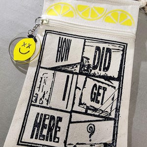 K&ouml;nnte beinhalten: Eine cremefarbene Canvas-Umh&auml;ngetasche mit Rei&szlig;verschluss und Kordelriemen. Die Tasche hat ein schwarzes Grafikdesign mit dem Text "HOW DID I GET HERE". Sie hat gelbe Zitronenscheiben-Designs und einen gelben Zitronenanh&auml;nger mit einem Smiley.