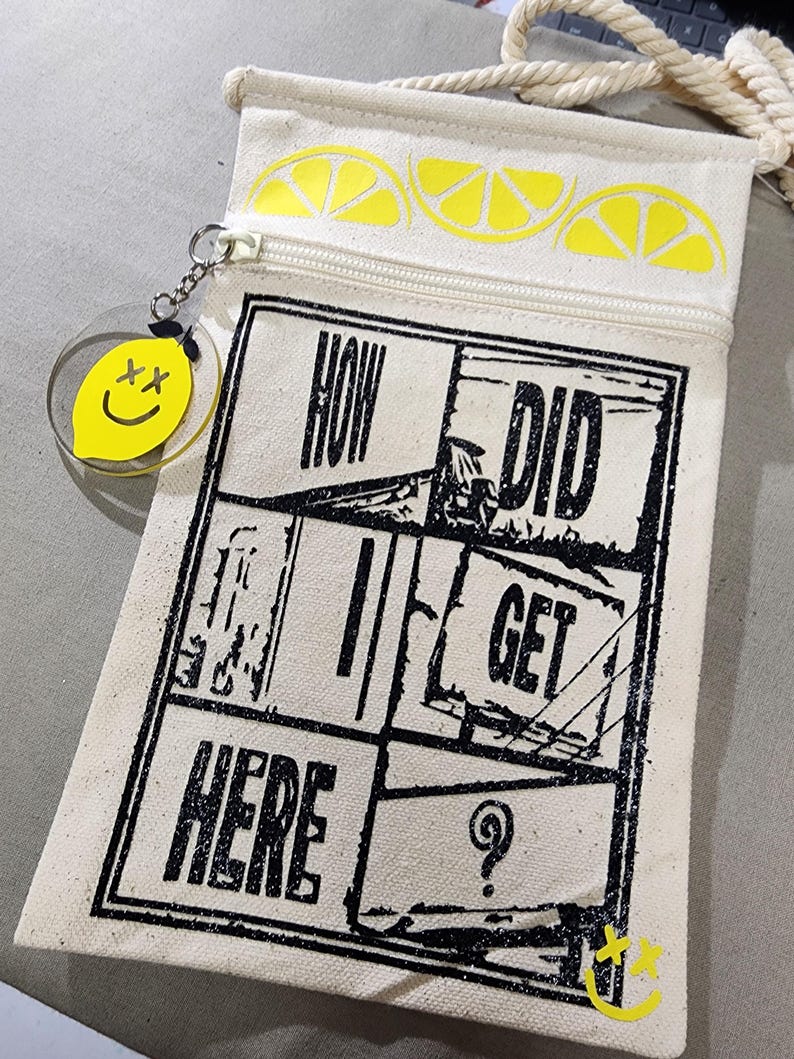 K&ouml;nnte beinhalten: Eine cremefarbene Canvas-Umh&auml;ngetasche mit Rei&szlig;verschluss und Kordelriemen. Die Tasche hat ein schwarzes Grafikdesign mit dem Text "HOW DID I GET HERE". Sie hat gelbe Zitronenscheiben-Designs und einen gelben Zitronenanh&auml;nger mit einem Smiley.